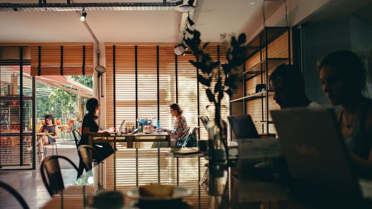 A woman working at a desk in an open-plan office, with colleagues at tables behind her.