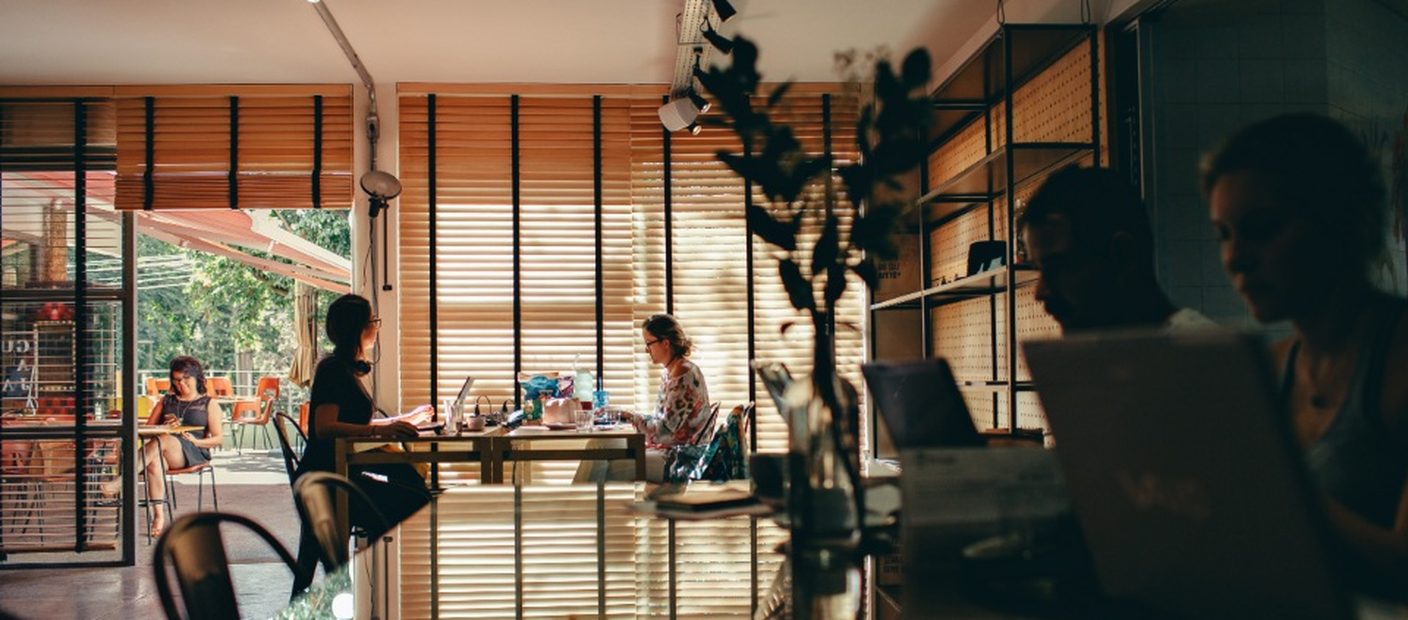 A woman working at a desk in an open-plan office, with colleagues at tables behind her.