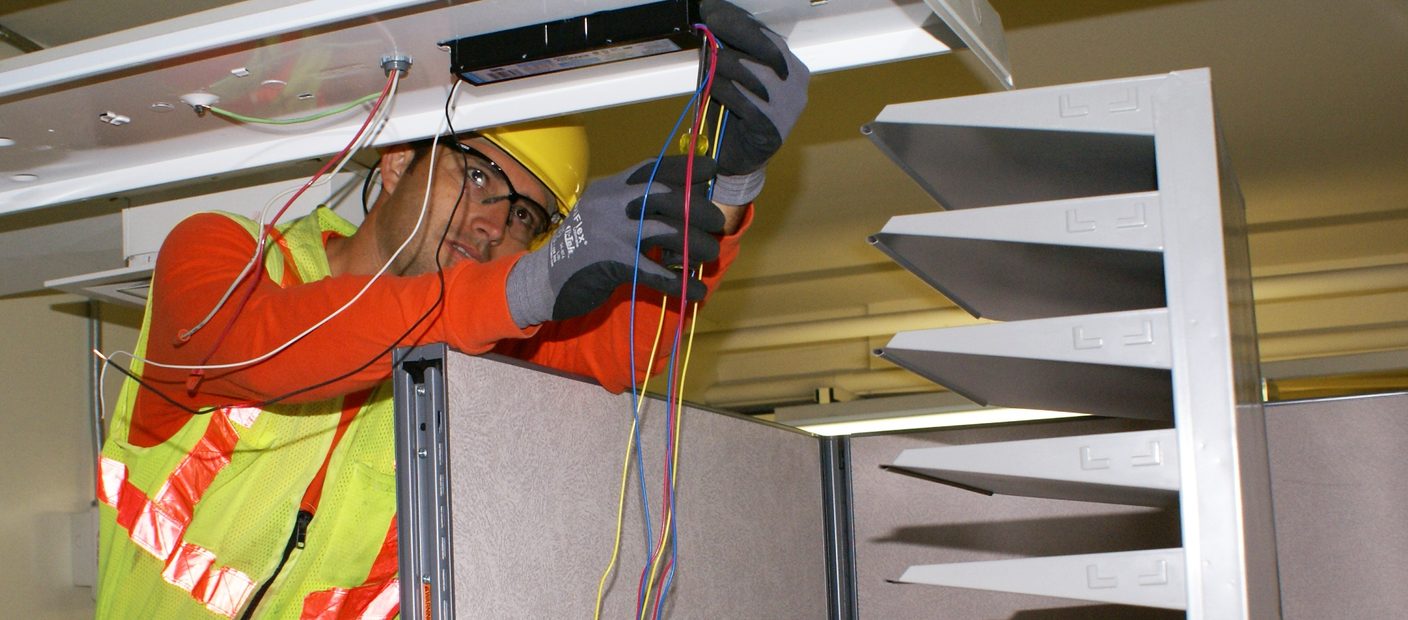 An electrician on a ladder rewiring a ceiling light fixture during a lighting retrofit.