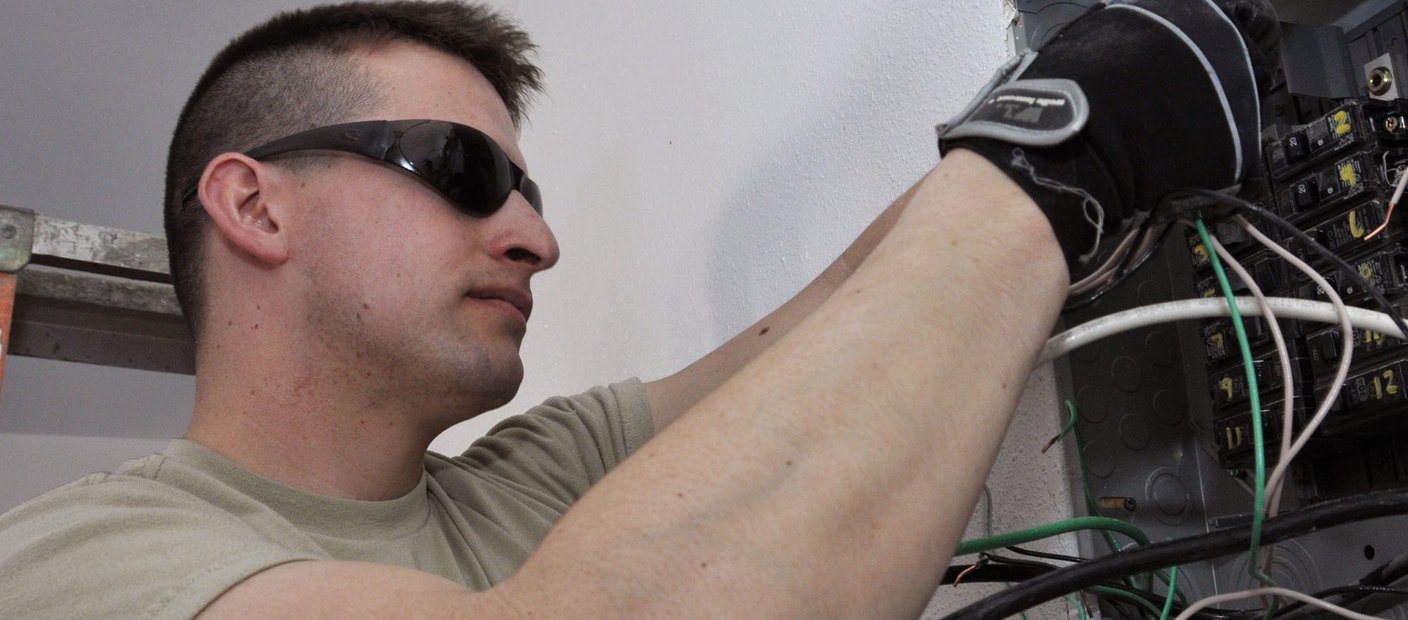 An electrician in work gloves connecting wiring inside an open electrical enclosure.