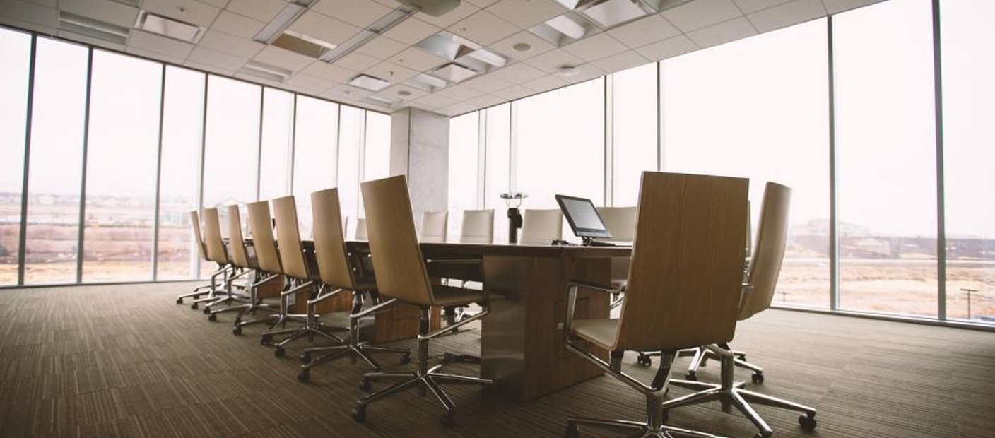 An empty boardroom with a long table and chairs, lit by a wall of windows.