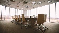An empty boardroom with a long table and chairs, lit by a wall of windows.