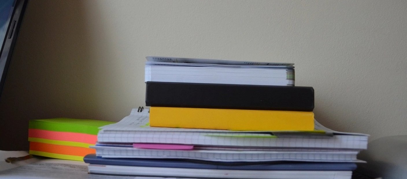 A tall stack of paper files and notebooks with coloured edges, unread.