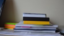 A tall stack of paper files and notebooks with coloured edges, unread.