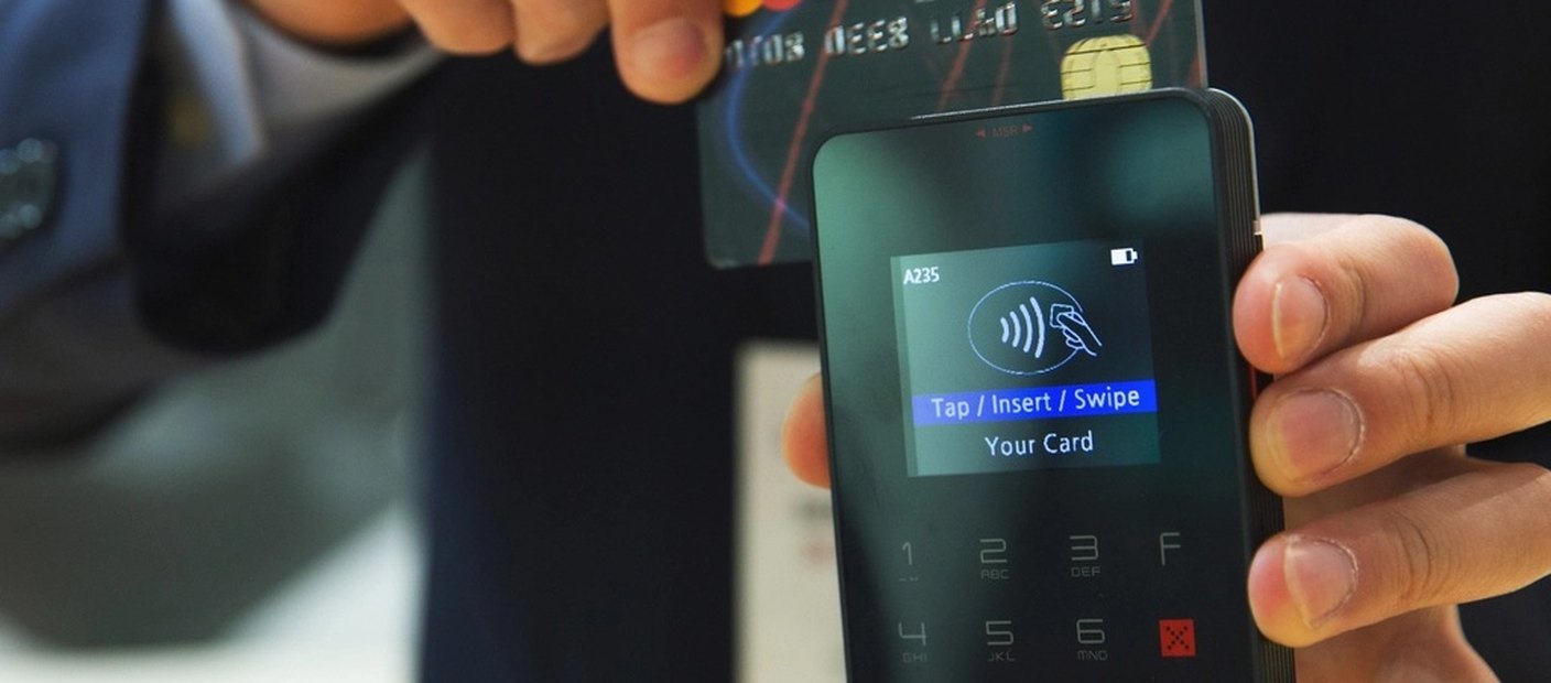 A hand holding a bank card against a hand-held contactless payment terminal.