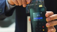 A hand holding a bank card against a hand-held contactless payment terminal.