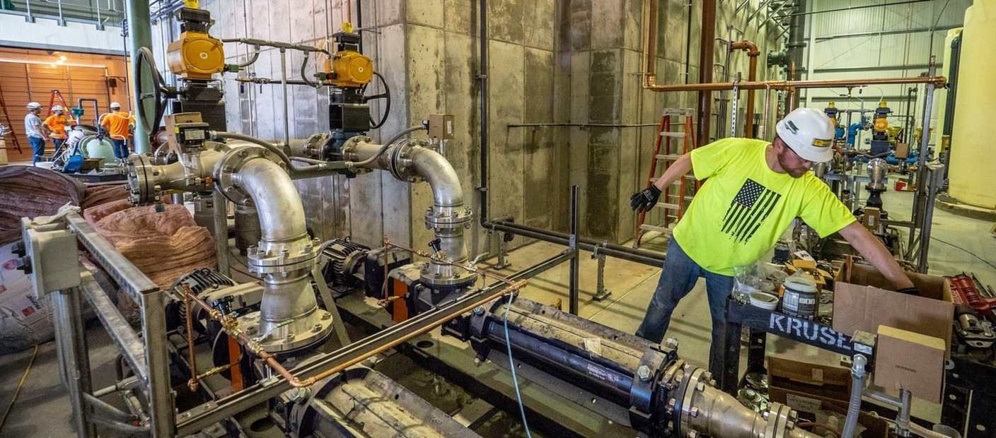A plumber in a hard hat and high-visibility vest working on large industrial pipework.