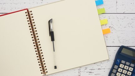 An open ruled notebook with a pen, coloured index tabs and a calculator on a white desk.
