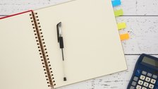 An open ruled notebook with a pen, coloured index tabs and a calculator on a white desk.