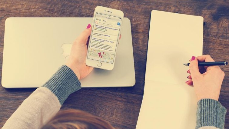 Hands writing on a blank notepad on a wooden desk while holding a phone.