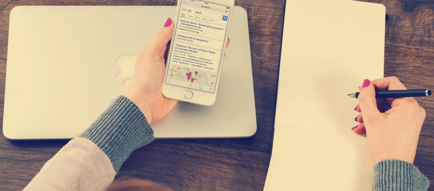 Hands writing on a blank notepad on a wooden desk while holding a phone.