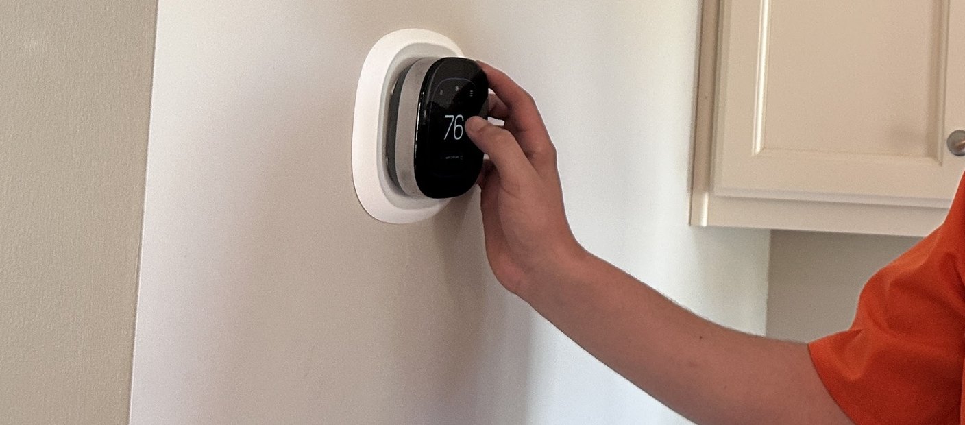A technician's hand setting a round wall thermostat inside a customer's home.