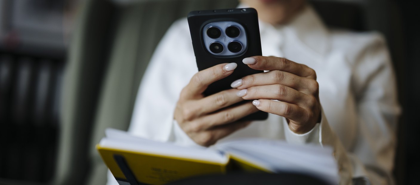 A seated person looking down at a smartphone while holding a notebook in the other hand.