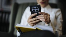 A seated person looking down at a smartphone while holding a notebook in the other hand.