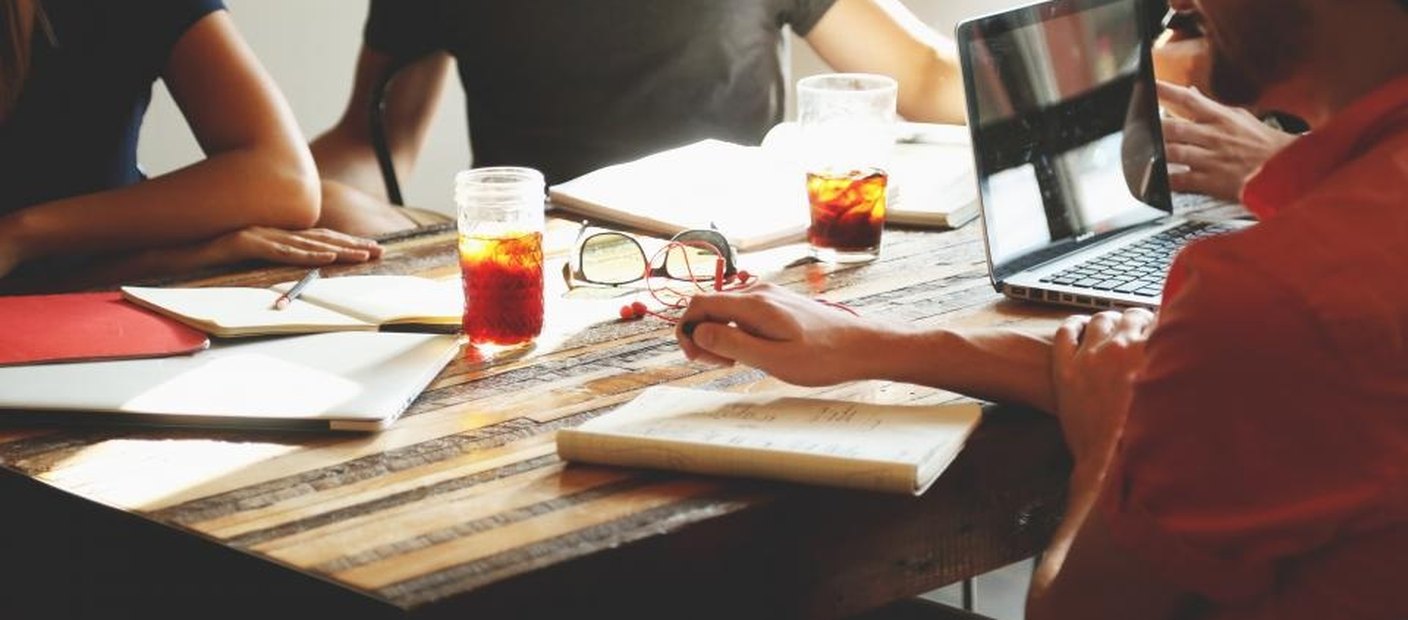 Several people around a table taking notes on paper and laptops during a meeting.