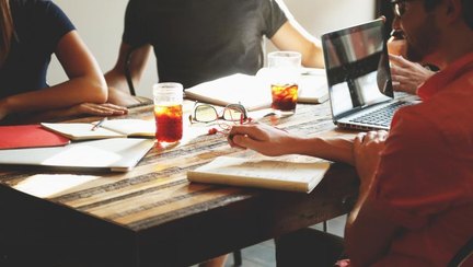 Several people around a table taking notes on paper and laptops during a meeting.