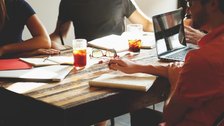 Several people around a table taking notes on paper and laptops during a meeting.